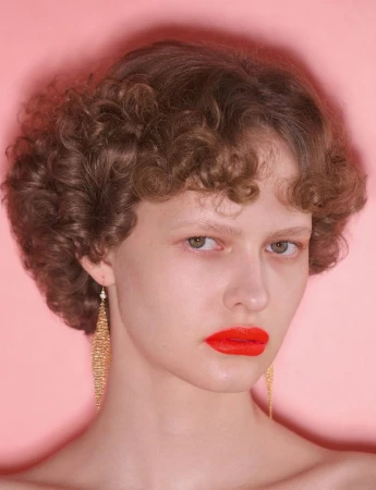 Close-up portrait of a woman with short curly brown hair, bright orange lipstick, and dangling gold mesh earrings against a pink background.