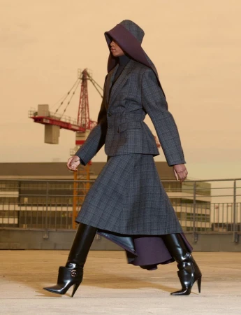 Model in plaid skirt suit and large hooded hat walking outdoors with a construction crane in the background