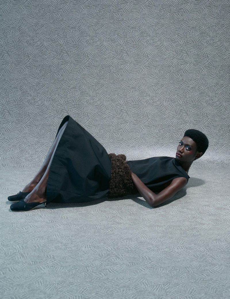 Model in dark sleeveless top and black skirt lying on a patterned textured backdrop for System Collections