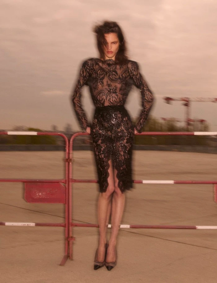 Model wearing a black lace dress leaning against a red and white barrier outdoors under an overcast sky.