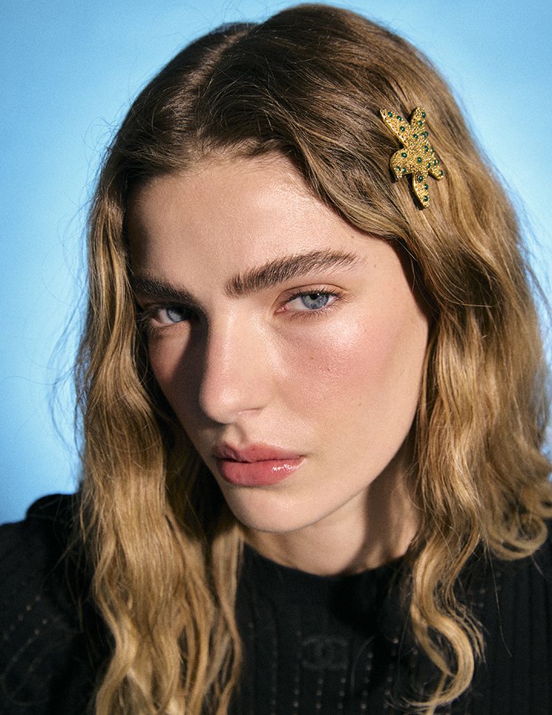 Close-up portrait of a woman with long wavy blonde hair wearing a gold star hair clip and black top against a bright blue background