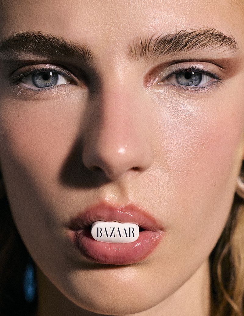 Close-up portrait of a woman holding a white pill with printed text between her lips for Harper's Bazaar Espana.