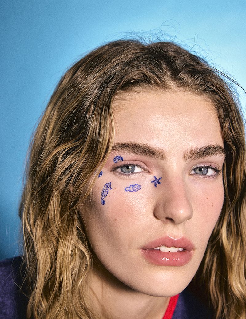 Close-up portrait of a woman with blue temporary tattoos on her face and shoulder-length brown hair against a bright blue background