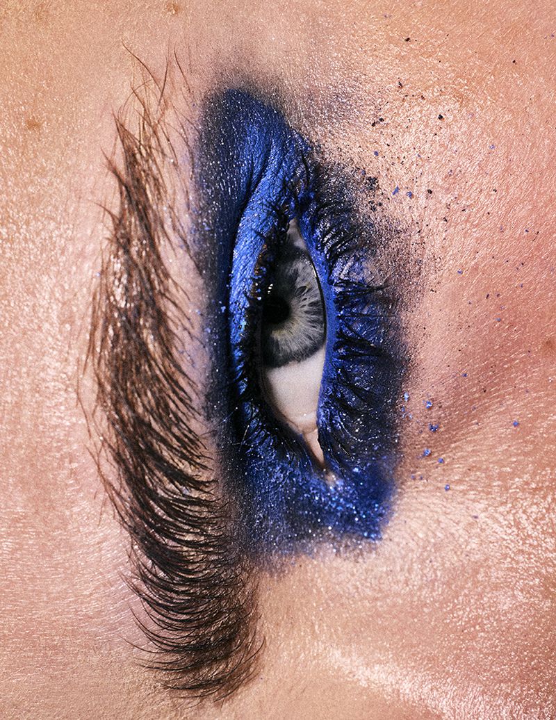 Extreme close-up macro view of a single human eye with dramatic royal blue eyeshadow, dark feathered eyelashes, and speckled blue pigment dust