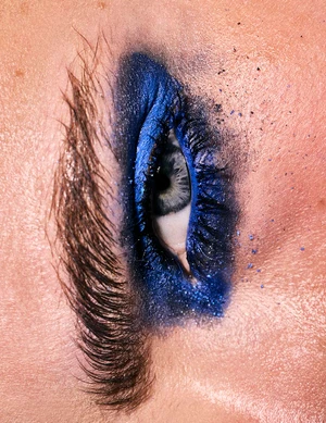 Extreme close-up macro view of a single human eye with dramatic royal blue eyeshadow, dark feathered eyelashes, and speckled blue pigment dust