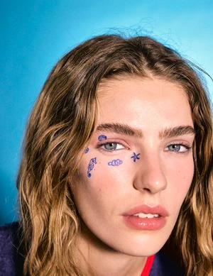 Close-up portrait of a woman with blue temporary tattoos on her face and shoulder-length brown hair against a bright blue background