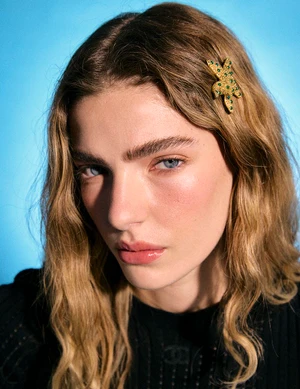Close-up portrait of a woman with long wavy blonde hair wearing a gold star hair clip and black top against a bright blue background