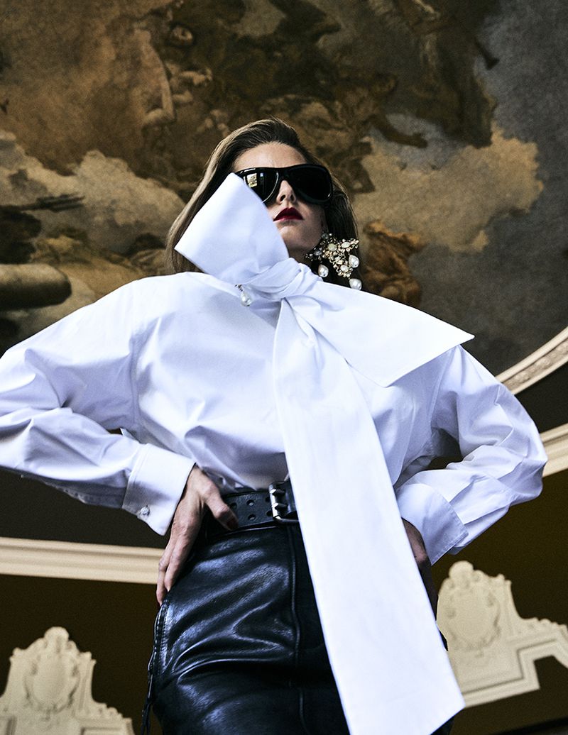 Model wearing a white blouse with a large bow, dark sunglasses, and black leather trousers beneath a painted ceiling fresco.