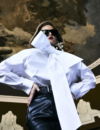 Model wearing a white blouse with a large bow, dark sunglasses, and black leather trousers beneath a painted ceiling fresco.