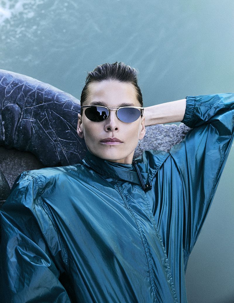 Model Hana Soukupova wearing metallic teal windbreaker and oval sunglasses resting near water for Harper's Bazaar España