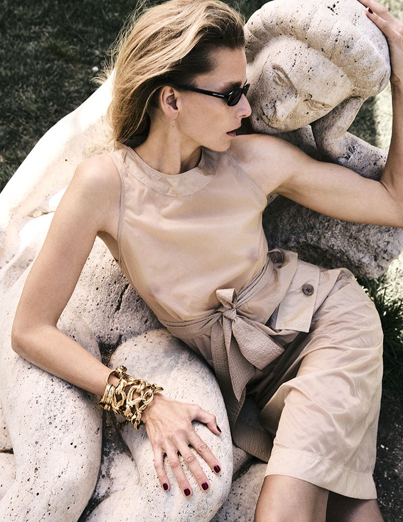 A blonde woman in beige clothing and sunglasses reclines against a white stone sculpture in an outdoor, sunlit setting for a fashion editorial.