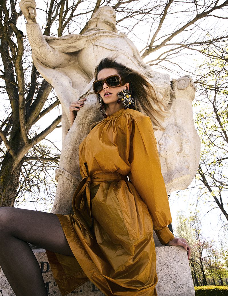Model wearing amber sunglasses and mustard coat posing in front of a stone sculpture under trees