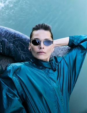 Model Hana Soukupova wearing metallic teal windbreaker and oval sunglasses resting near water for Harper's Bazaar España
