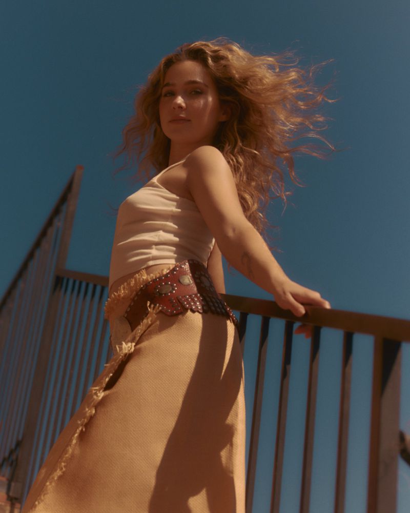 Haley Lu Richardson leaning against a railing against a clear blue sky, wearing a beige tank top and studded belt.