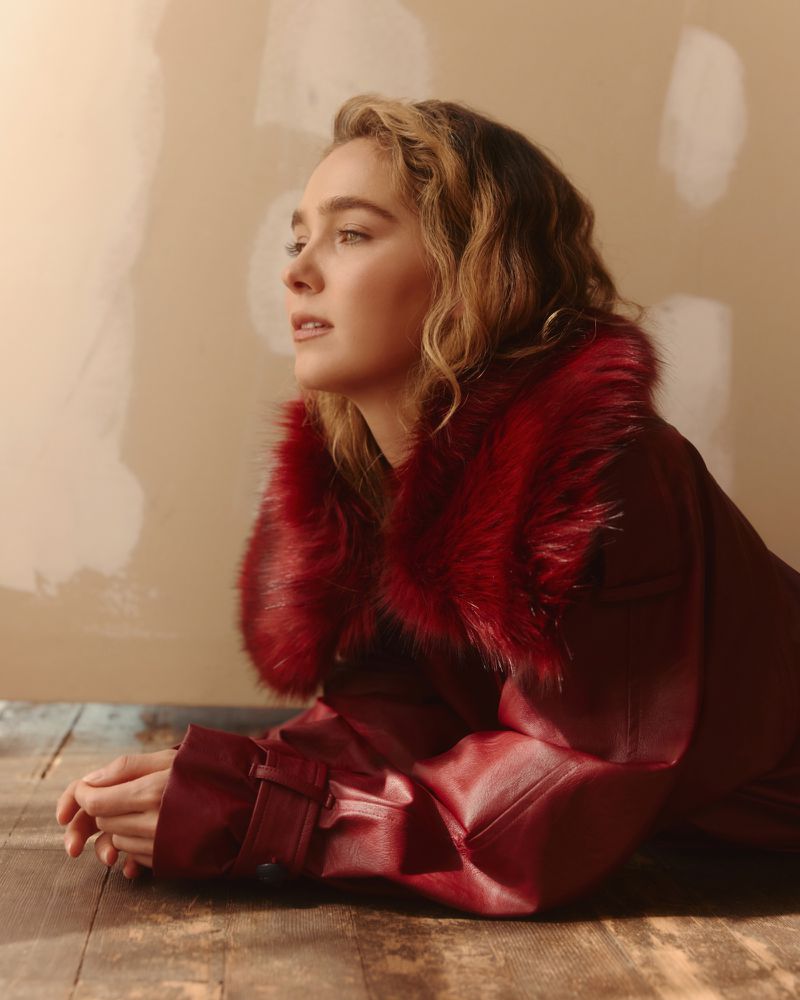 Haley Lu Richardson wearing a red coat with a furry collar, posed on a wooden floor against a neutral wall.