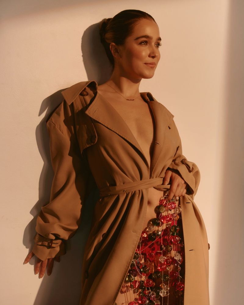 Haley Lu Richardson wearing a camel trench coat and beaded skirt against a plain wall in warm light for Grumpy Magazine