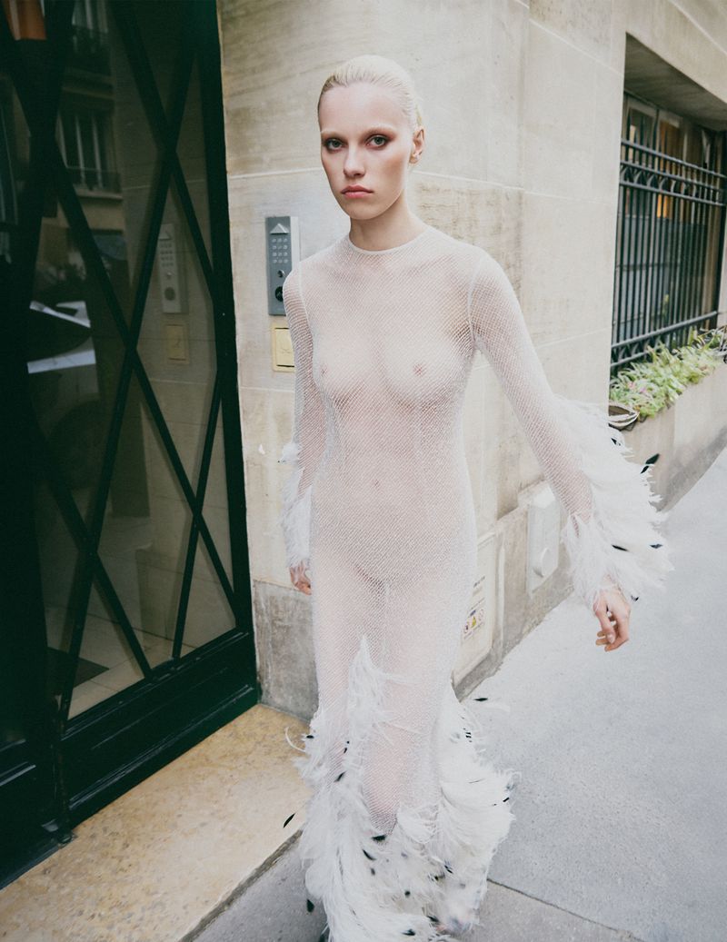 Model in a sheer white feathered dress walking on an outdoor stone sidewalk past a building entrance