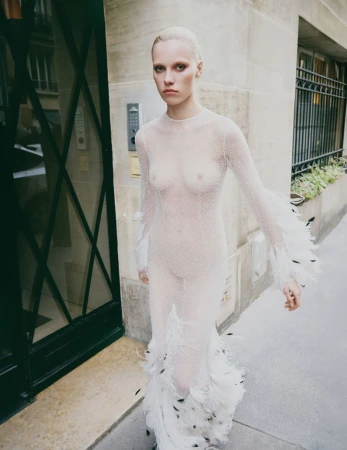 Model in a sheer white feathered dress walking on an outdoor stone sidewalk past a building entrance