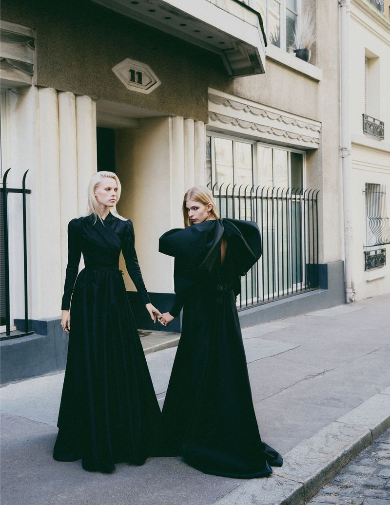 Two female models in long black dresses standing outside an apartment building entrance in a street editorial for Flaunt.