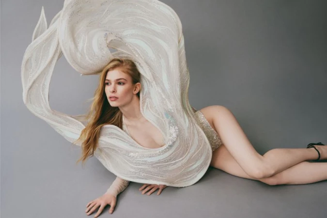 Model lying on a grey studio backdrop wearing a flowing sculptural white sheer garment with metallic detailing