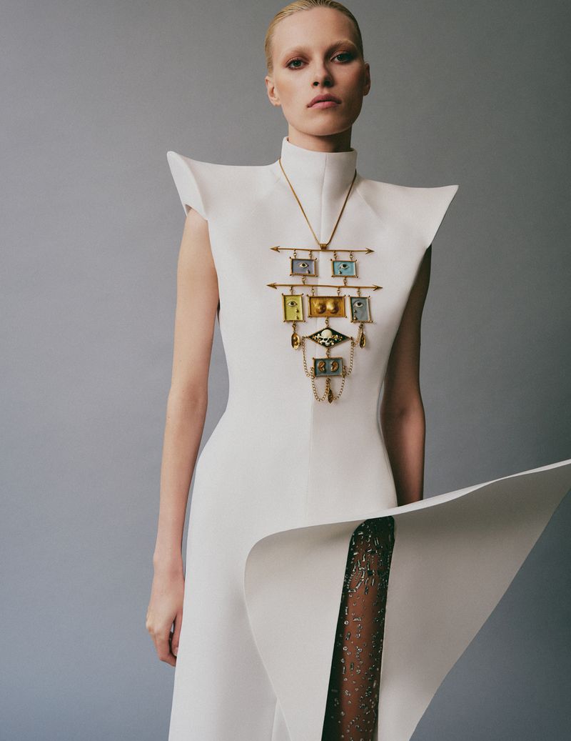 Model in architectural white dress wearing an ornate pendant necklace against a grey studio backdrop