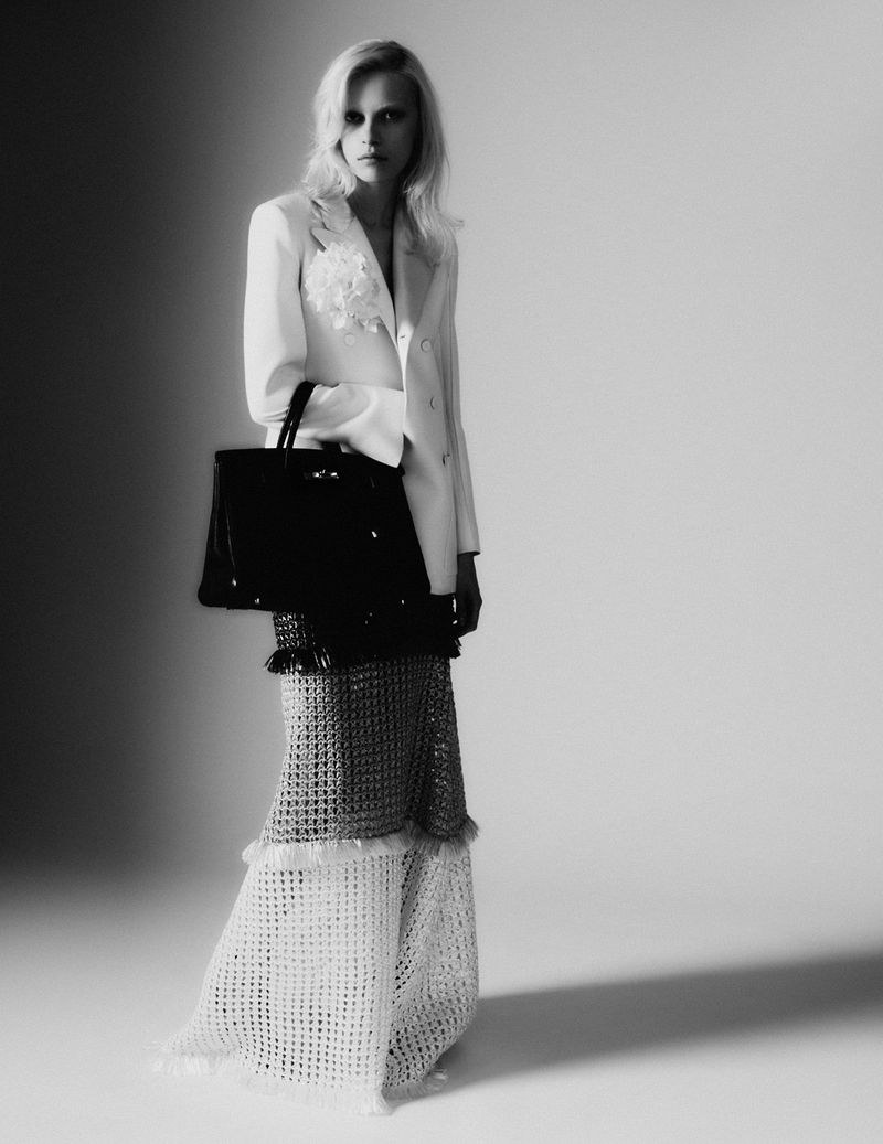 Model in white blazer and patterned maxi skirt holding a black handbag against a grey studio backdrop.