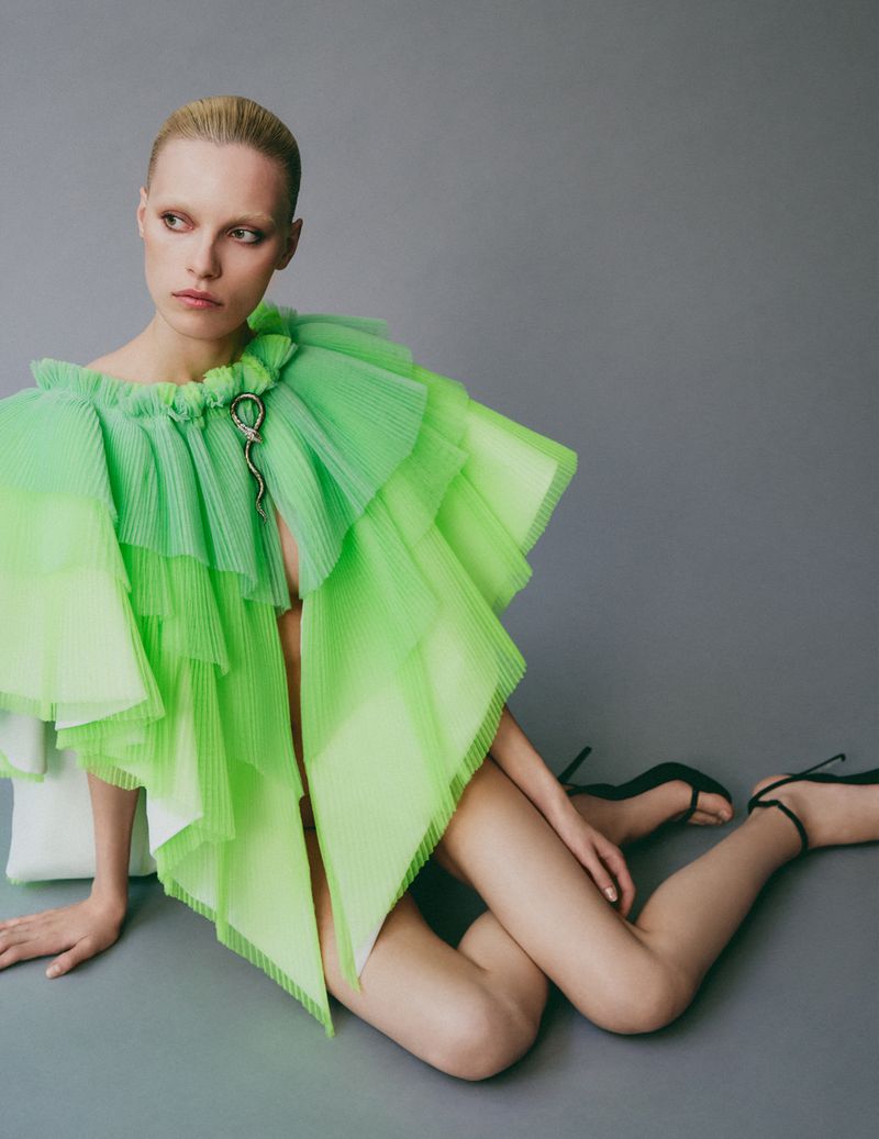 Model with slicked-back blonde hair wearing a bright green ruffled cape top against a neutral grey studio backdrop.