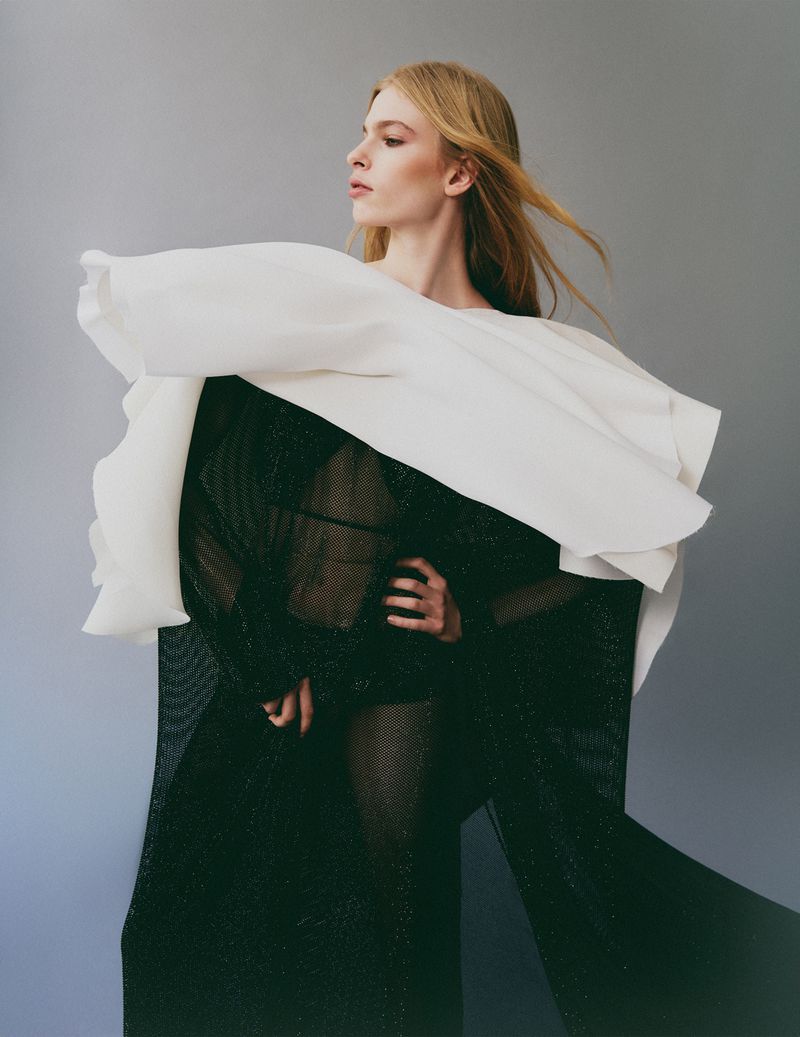 Model wearing a structured white overlay and black sheer mesh garment against a grey gradient studio background.