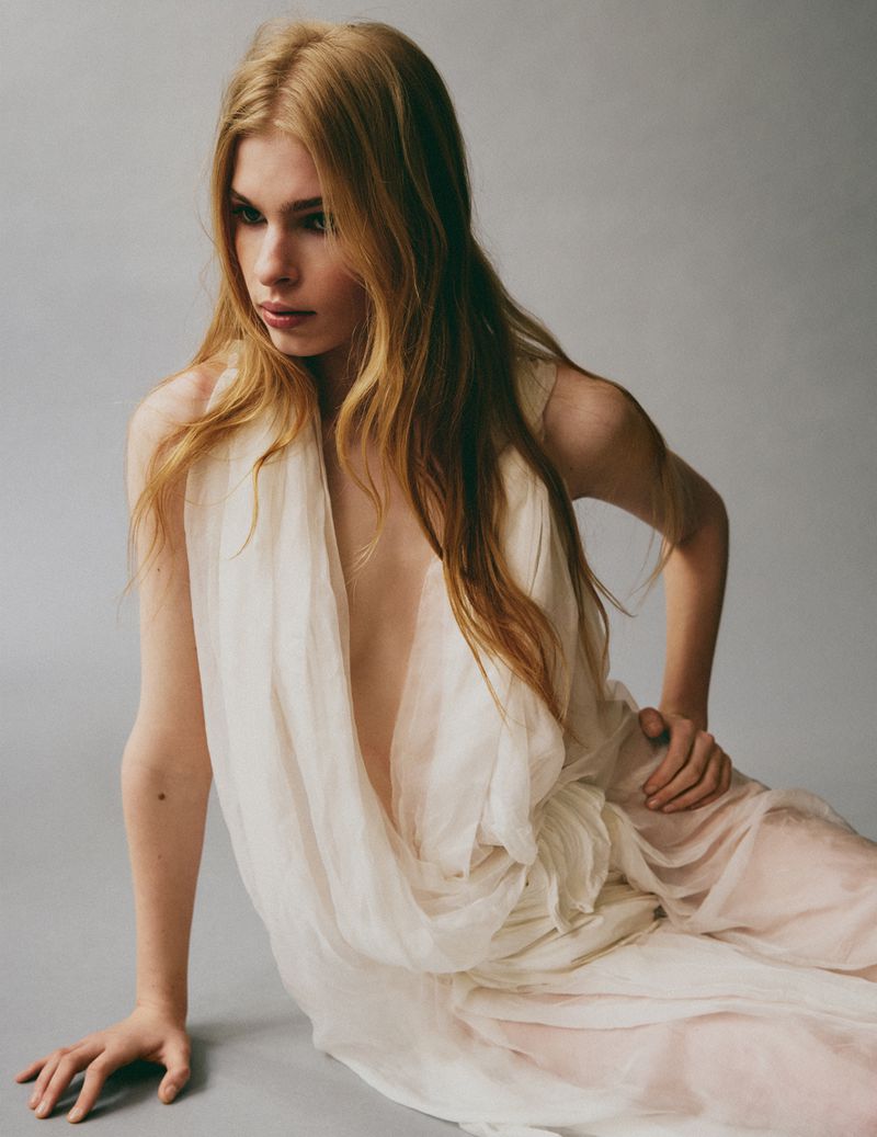 Model with long blonde hair wearing a white draped dress seated against a grey studio seamless background