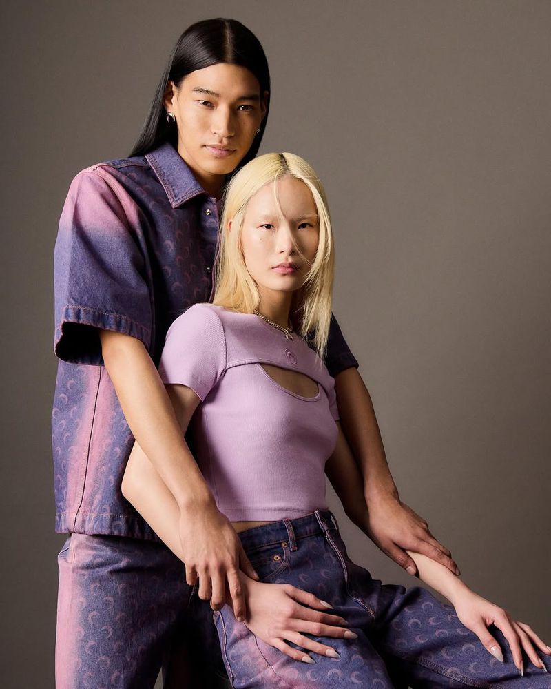 Two fashion models, a man and a woman, pose indoors wearing matching purple-and-pink outfits with crescent moon patterns against a grey background.