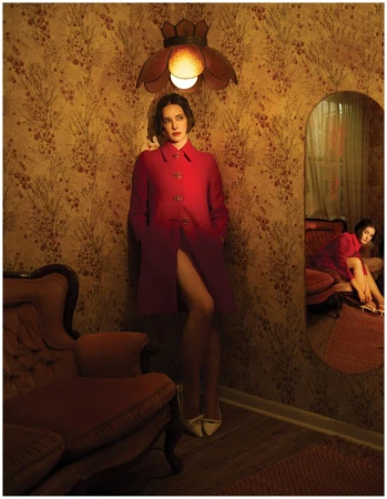 Hari Nef in a bright red coat standing against patterned wallpaper in a vintage-styled interior room.