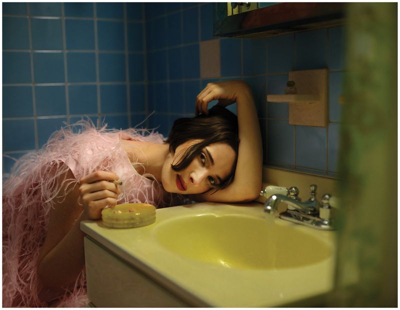 Hari Nef wearing a pink feathered dress leaning on a yellow bathroom sink against blue tiled walls