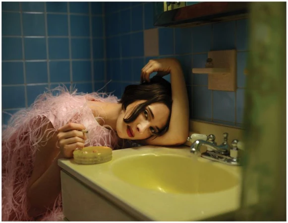 Hari Nef wearing a pink feathered dress leaning on a yellow bathroom sink against blue tiled walls