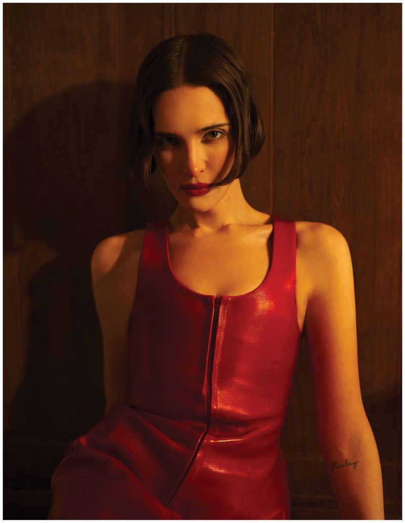 Hari Nef wearing a red leather dress against a dark wood wall with warm low-key lighting for Schön Magazine.