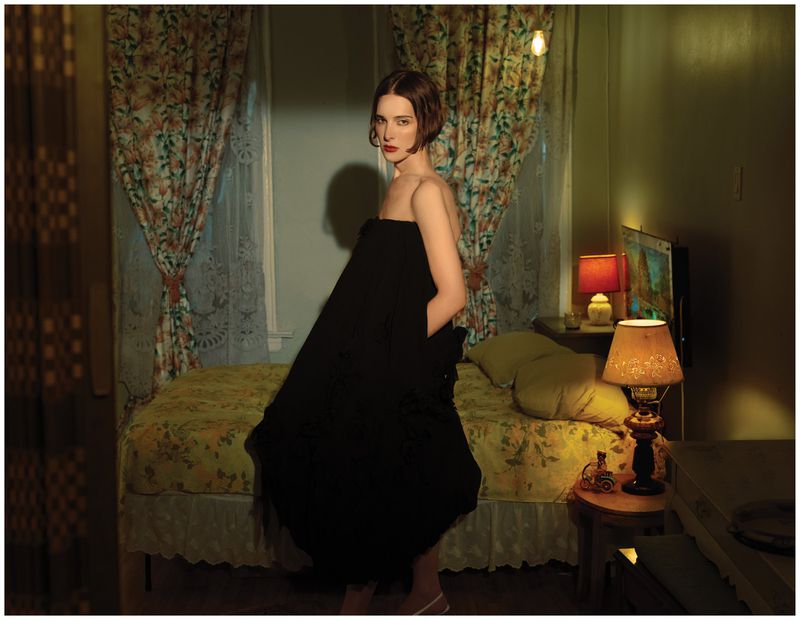 Hari Nef wearing a black strapless dress in a warmly lit bedroom with floral curtains