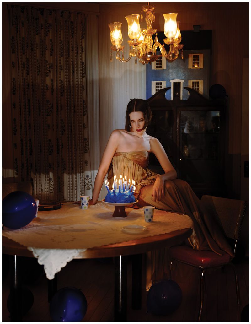 Hari Nef in a gold strapless dress sitting on a table with a lit birthday cake in a dim room.