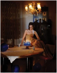 Hari Nef in a gold strapless dress sitting on a table with a lit birthday cake in a dim room.