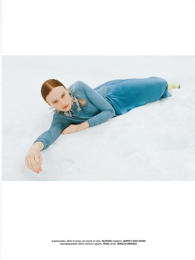 Model lying on a white seamless backdrop wearing a long-sleeved blue dress and feathered collar necklace.