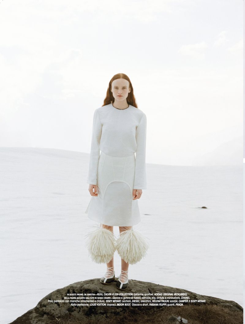 Model standing on a dark rock formation against a pale, overcast landscape wearing white clothing