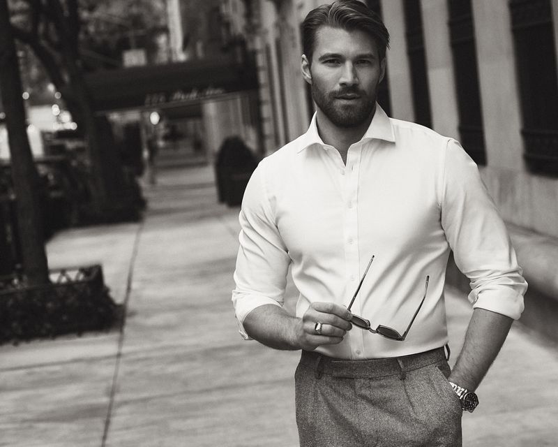 Monochrome portrait of an adult man in a button-down shirt and dress pants, holding sunglasses on a city sidewalk.