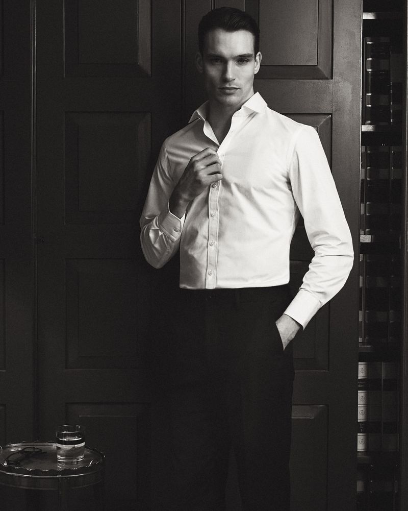 Man wearing a white button-down shirt and dark trousers standing indoors against a dark wooden paneled wall