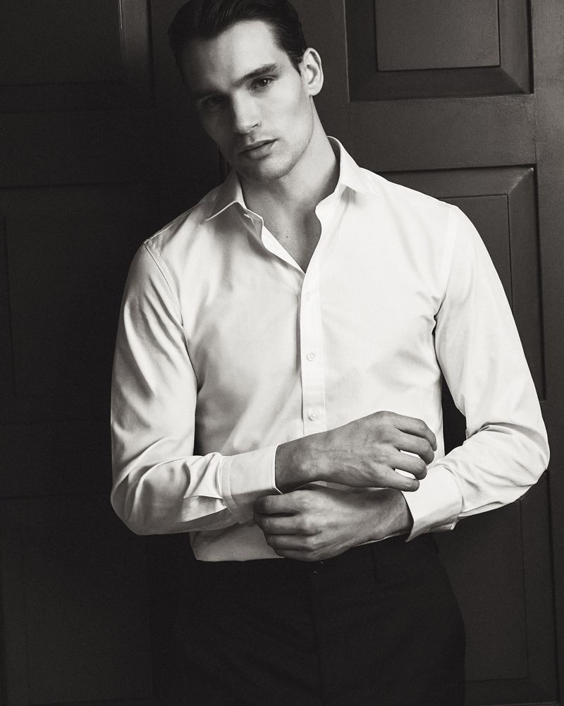 Man wearing a white dress shirt buttoned at the collar with cuffs adjusted against a dark paneled wooden wall.