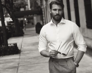 Monochrome portrait of a bearded man wearing a white shirt and trousers holding sunglasses on a city sidewalk