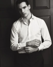 Man wearing a white dress shirt buttoned at the collar with cuffs adjusted against a dark paneled wooden wall.