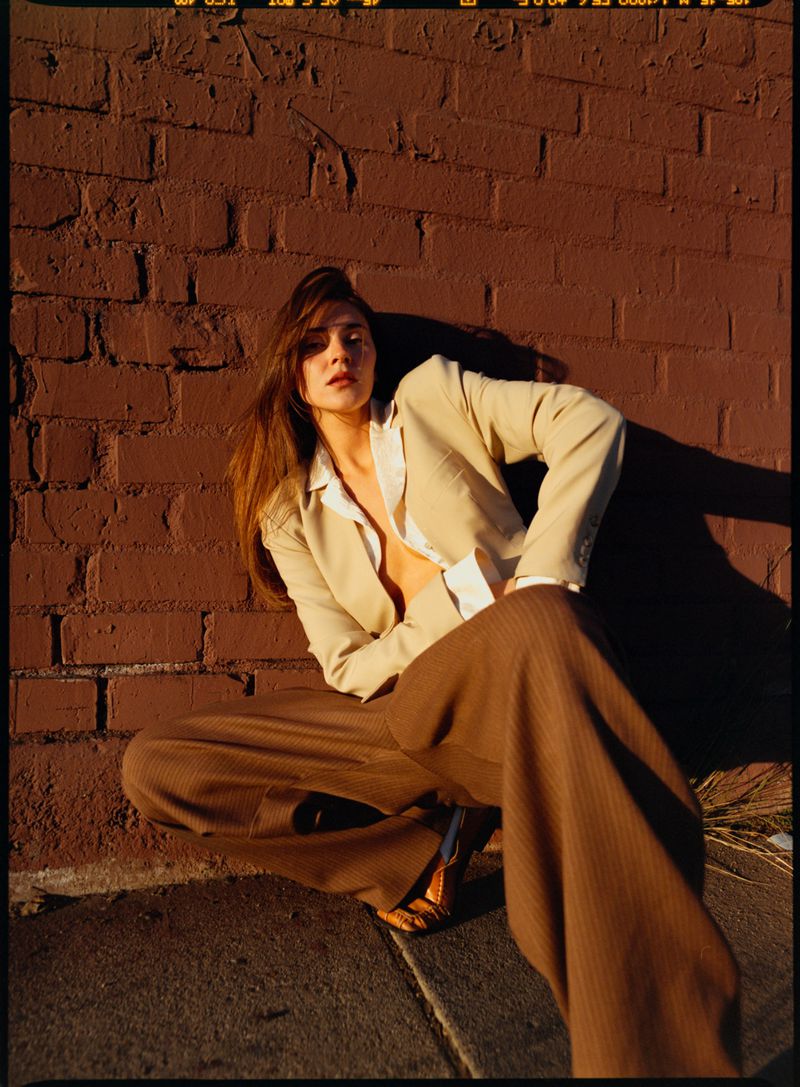 Model Stefanie Giesinger squatting in a beige blazer and brown trousers against a dark red brick wall