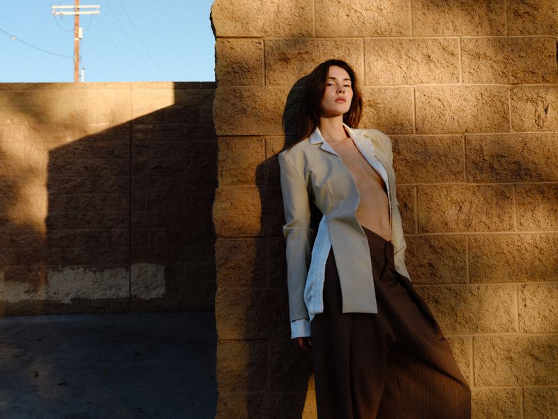 Model Stefanie Giesinger leaning against a textured brown brick wall wearing a structured beige jacket and dark trousers.