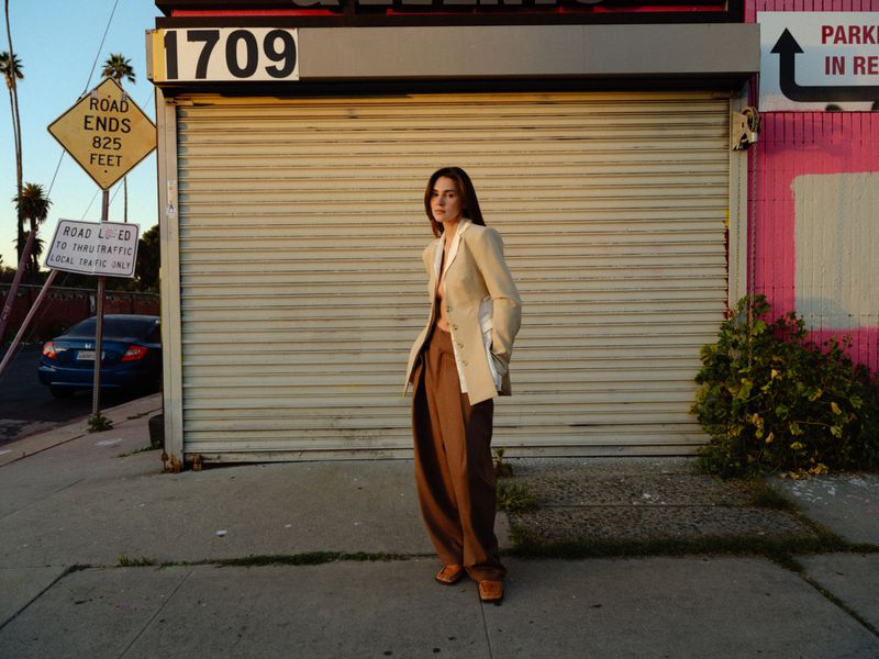 Model Stefanie Giesinger standing in a beige blazer and brown trousers on a street in front of a closed metal shutter storefront.