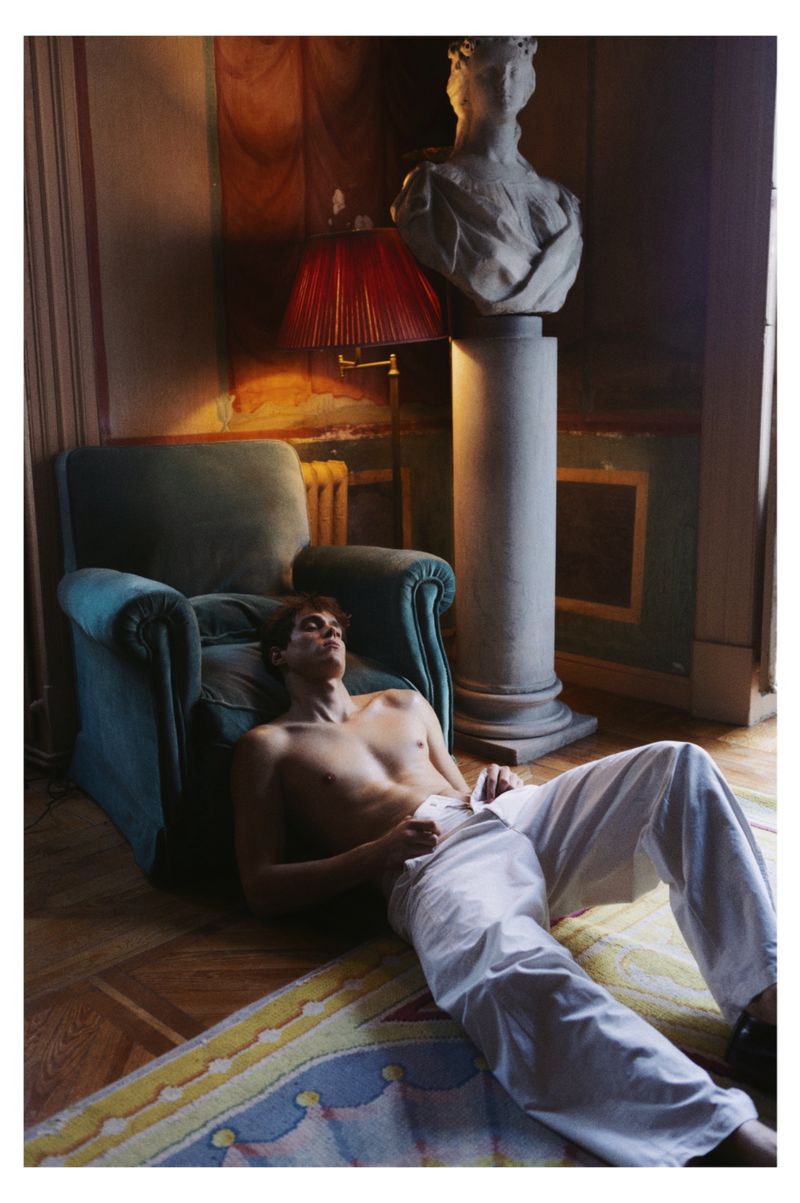 Shirtless man sitting on parquet floor leaning against a teal armchair with a classical bust and red lamp behind him.