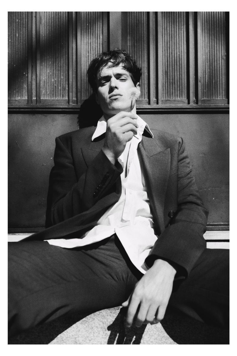 Monochrome portrait of a man in a tailored suit jacket holding a dandelion flower against a dark paneled wall.
