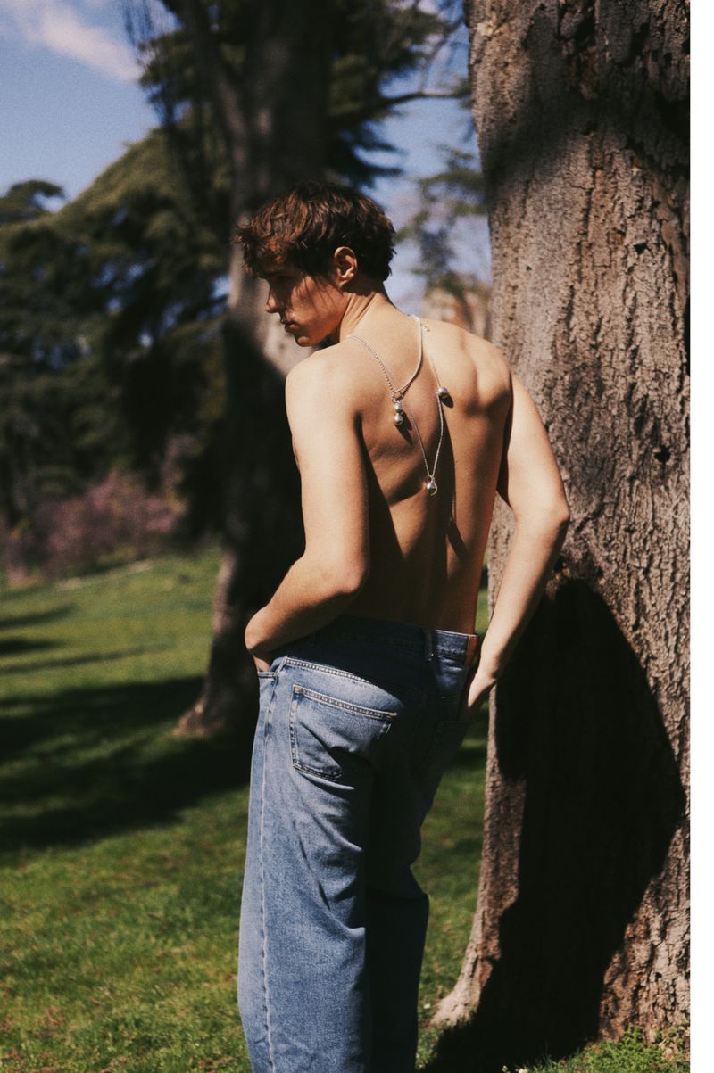 Shirtless man wearing blue jeans and silver pendant necklace leaning against tree trunk in a park